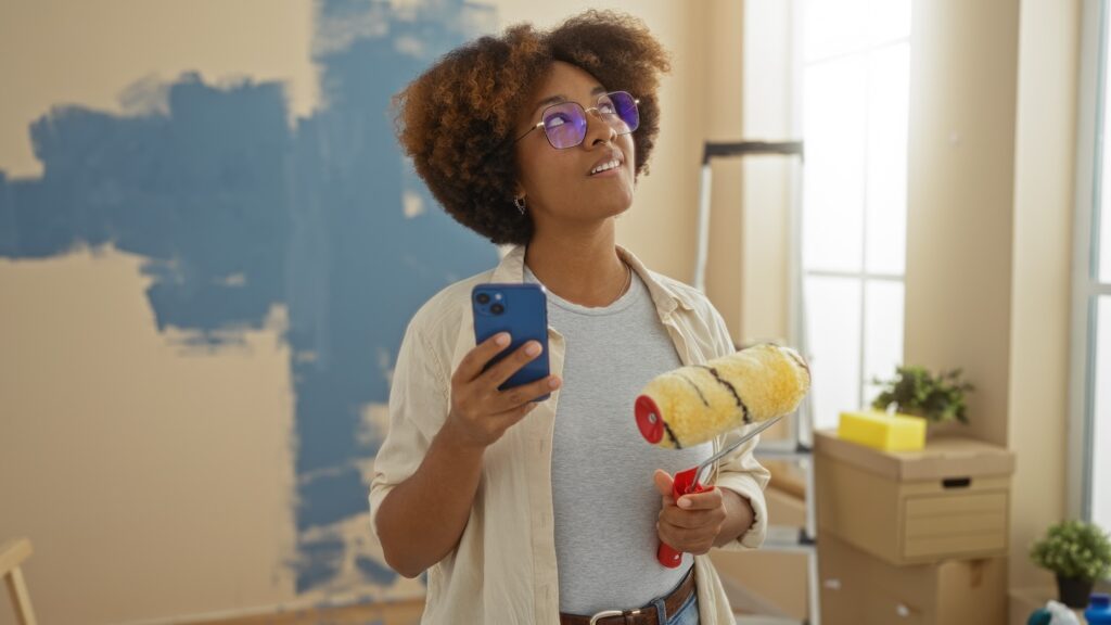 A homeowner stands in a sunlit, freshly painted blue room—phone in one hand, paint roller in the other—actively searching Google for reliable home services. Boxes and supplies are visible in the background, highlighting the need for trusted professionals during home projects.