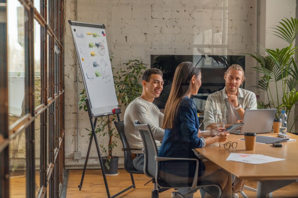 Three professionals meet in a modern office to discuss outsourced marketing strategies. They collaborate over laptops and documents, exchanging ideas in a bright space with large windows and greenery. A flip chart with diagrams highlights the strategic planning underway.