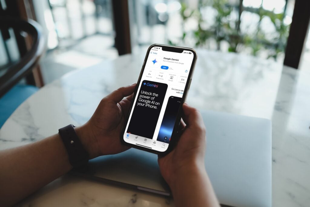 A person holds an iPhone open to the Google Gemini app page on the App Store, showcasing features like Generative Engine Optimization (GEO) designed for home service businesses. In the background, a blurred laptop sits on a marble table near a window with greenery outside.
