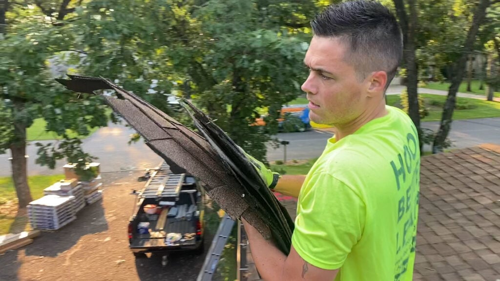 A roofing professional in a bright yellow shirt stands on a roof, confidently holding several shingles—demonstrating our deep understanding of the roofing industry. In the background, trees and a work truck loaded with roofing materials reinforce our firsthand knowledge of your business.