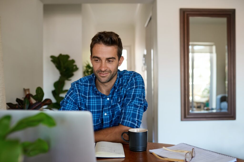 A man in a blue plaid shirt sits at a table with his laptop, notebook, and coffee mug. He smiles at his screen as he explores emerging AI marketing trends for home service businesses in 2025, surrounded by papers, plants, and a mirror.