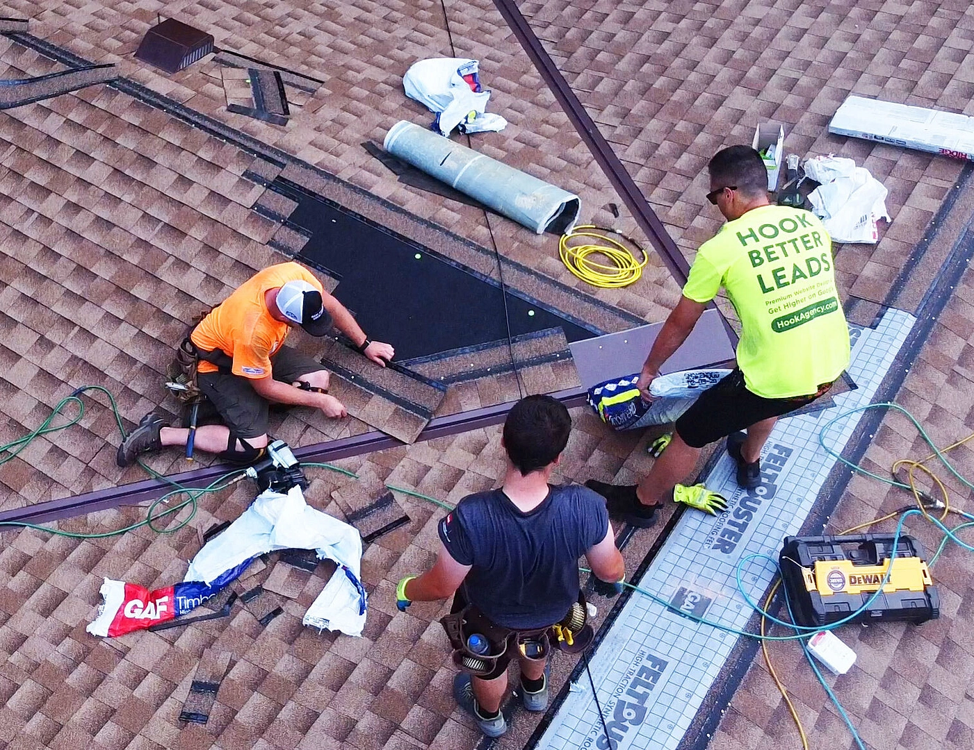 Three workers in safety gear install or repair a brown shingle roof, using tools, cables, and green roofing materials. One wears a shirt with the slogan "HOOK BETTER LEADS.