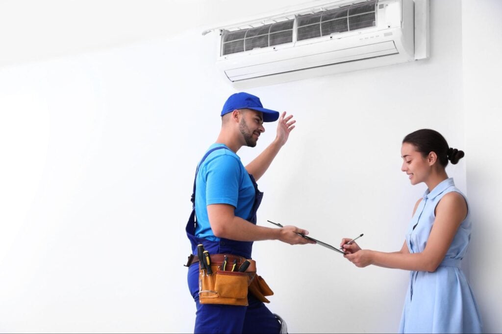 HVAC technician in blue uniform gestures at wall AC as client signs clipboard, demonstrating key tasks of the journeyman role.