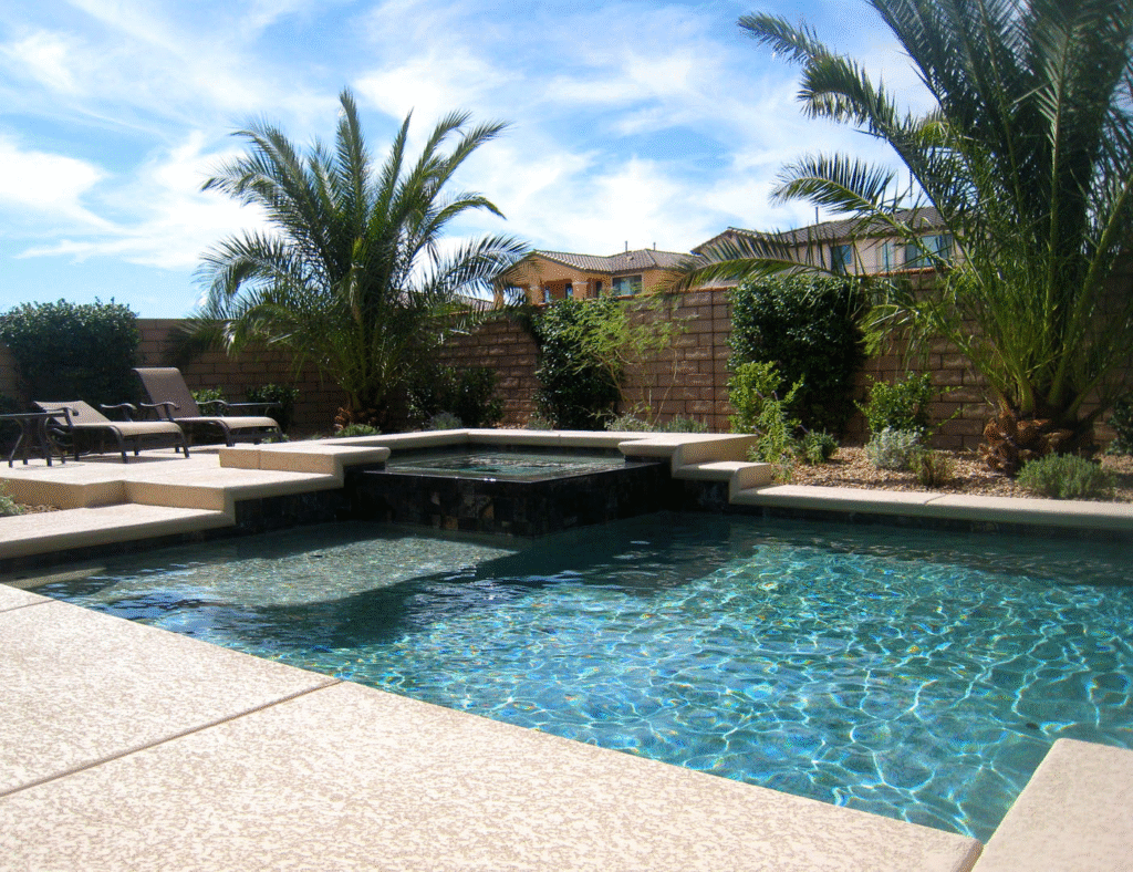 Premium U.S.-built pool with raised hot tub, stone deck, lounge chairs, palm trees, and privacy fence in a sunny backyard.