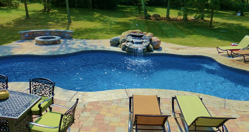 Professionally designed backyard pool with clear water, waterfall, fire pit, lounge chairs, dining table, patio, and greenery.