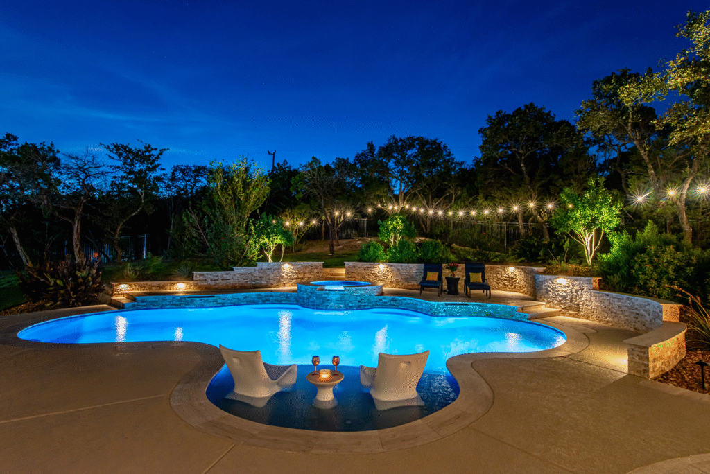 Backyard pool glows blue at night, with lounge chairs, drinks table, expert stonework and lush landscaping—by Hook Agency.