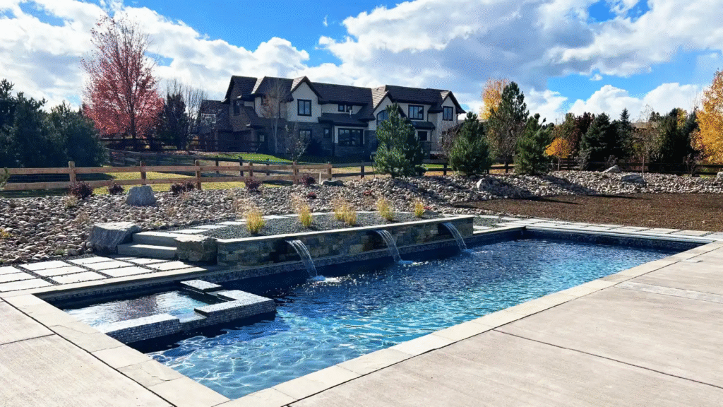 Rectangular pool with three waterfalls and hot tub by leading installers, in a landscaped backyard with trees and large house.