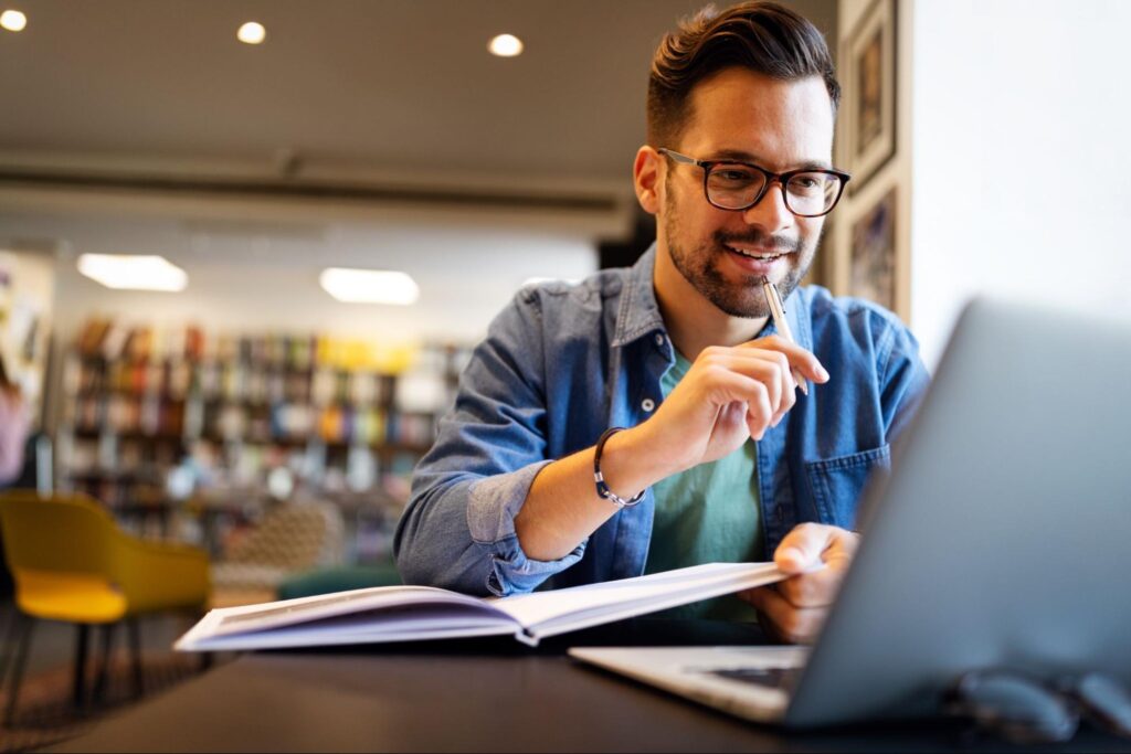 Professional man with glasses works at a laptop, pen in hand, notebook open, in a modern office or cafe settingāfocused and productive.