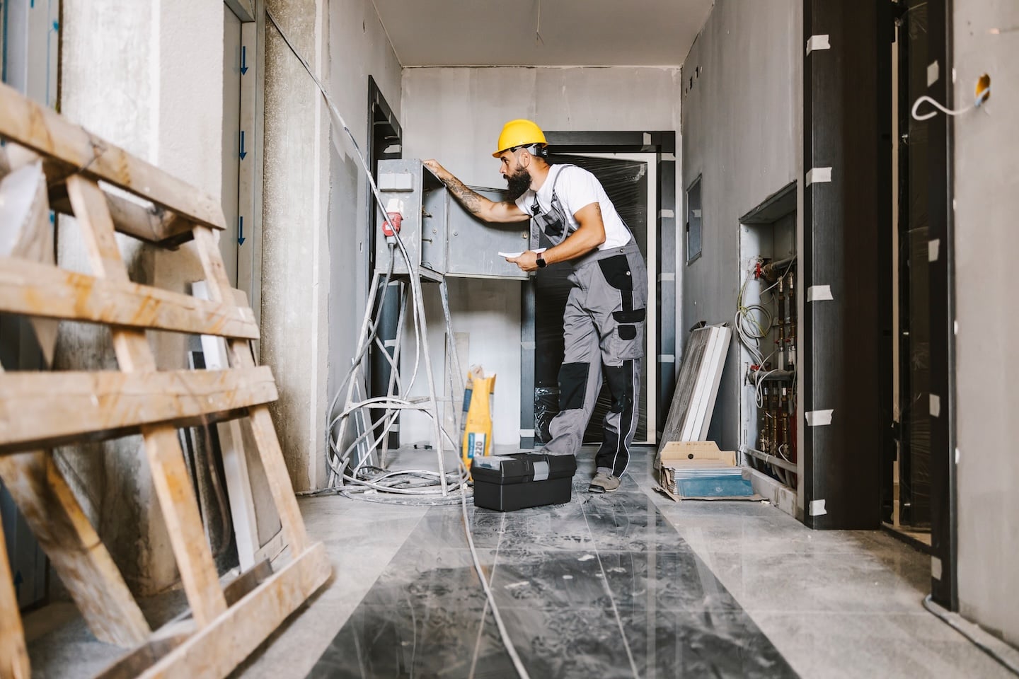 Construction worker in yellow hard hat inspects wiring and HVAC in renovation, clipboard in hand near exposed walls and tools.