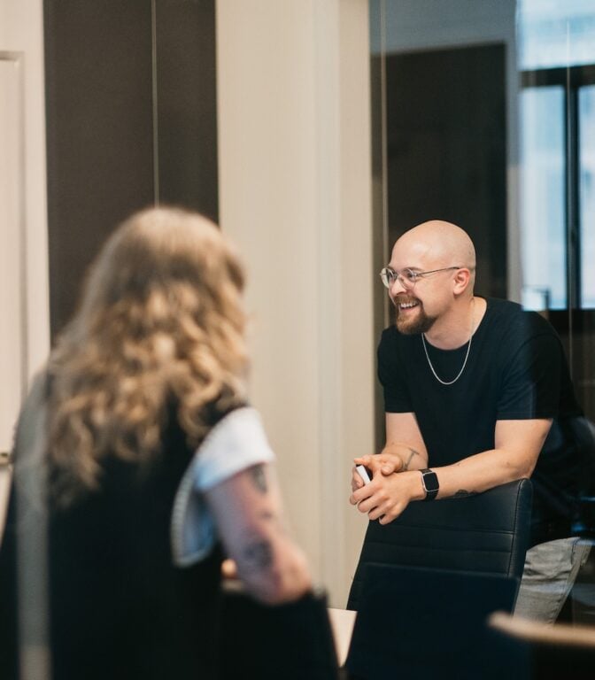 Smiling bald man with glasses consults on HVAC LSA management with long-haired colleague in office—engaged, expert discussion.