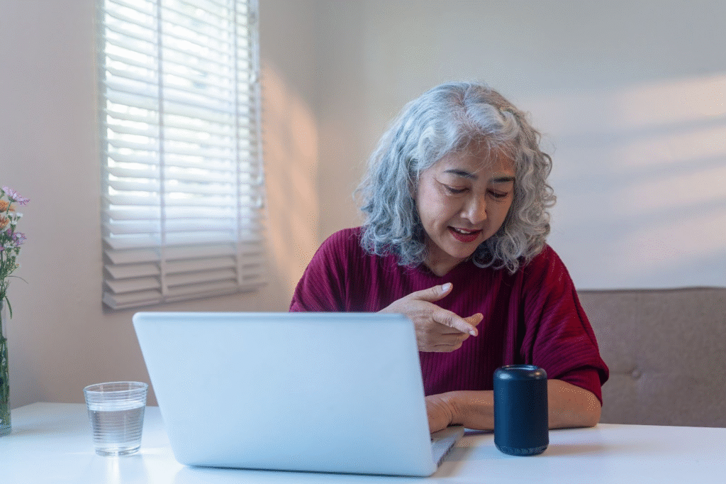 Older woman uses voice search on a smart speaker at her desk with laptop and water, illustrating digital accessibility.