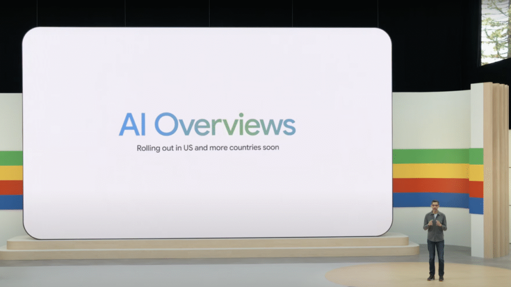 Presenter at a tech event with a screen announcing AI Overviews launching soon in more countries; vibrant stripes in background.