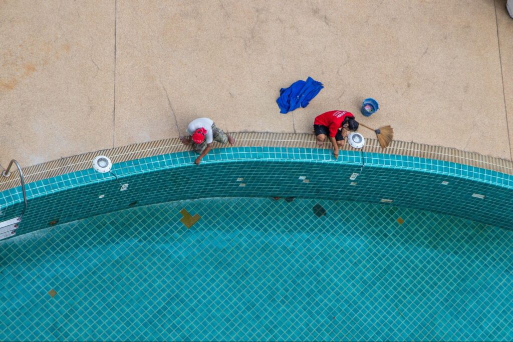 Two people clean a pool edge with brooms; cleaning supplies and a blue towel sit nearby—ideal visual for Pool Installer Tips.