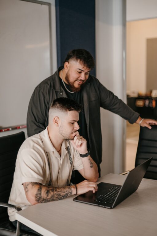 Two men collaborate at a table; one reviews an SEO dashboard on a laptop as the other stands beside him, analyzing the data.