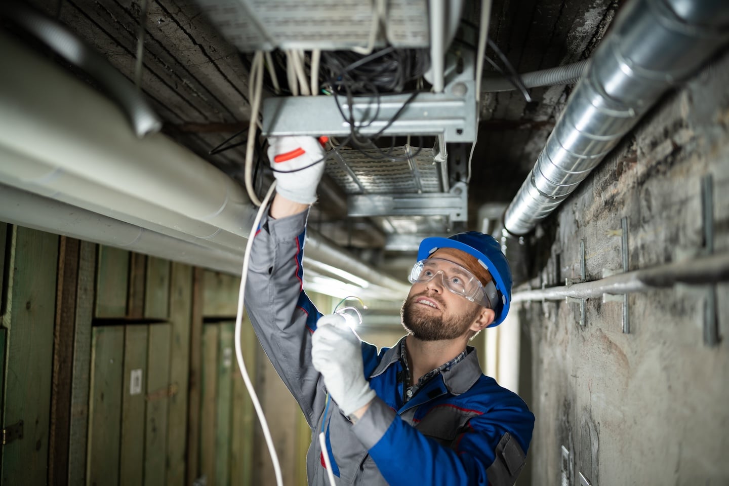 AEO worker in blue helmet and goggles safely installs or repairs ceiling electrical wiring, using tools and handling overhead cables.