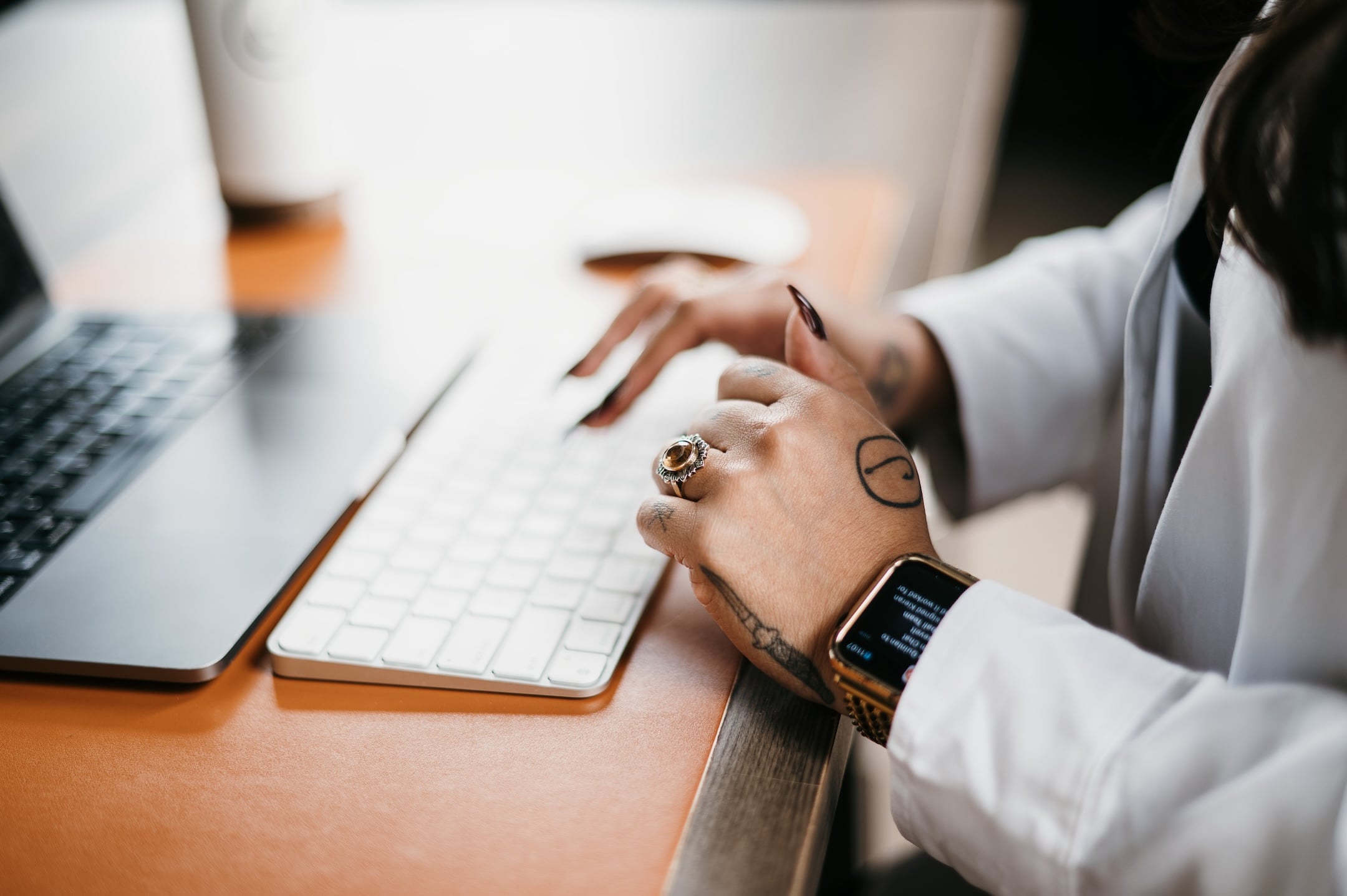 A professional types on a white keyboard beside a laptop, wearing rings and a smartwatch, with a blurred cup in the background.