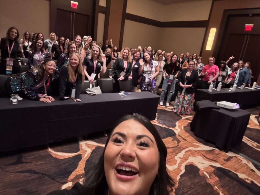 A woman takes a selfie with a large group of smiling tradeswomen in a conference room, with tables and badges visible.