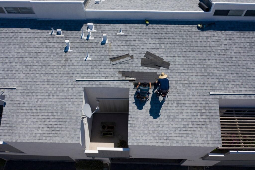Roofers repair a gray shingle roof with tools, beside vents and courtyard—perfect for showcasing quality roofing work by Hook Agency.