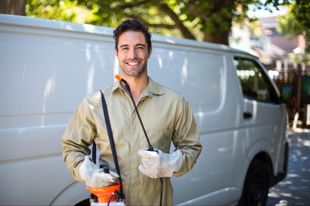 Professional pest control technician in uniform and gloves stands by branded van, ready to serve. Sunlit trees in the background.