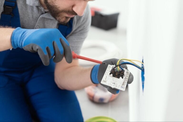 Electrician in blue gloves and overalls uses screwdriver on wall outlet, showing expert service featured by Hook Agency.