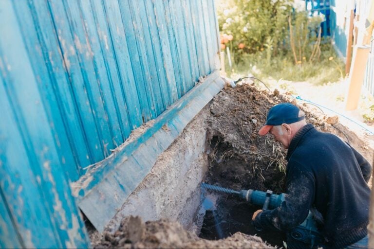 A technician drills into a blue building's foundation near an outdoor trench—demonstrating Hook Agency's expert repair services.