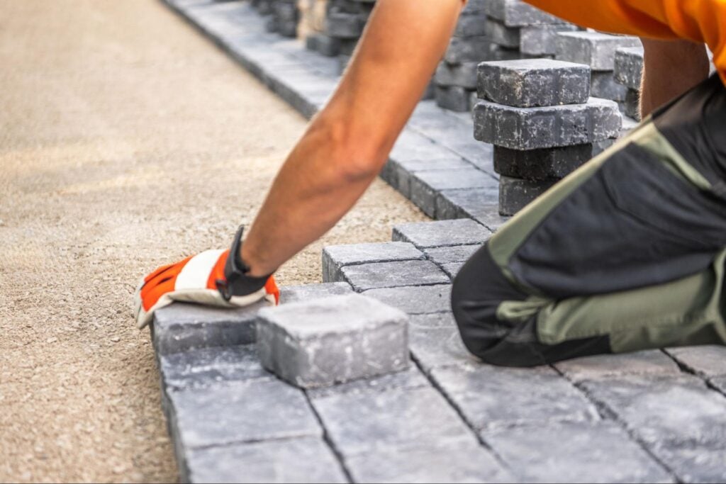 Hardscape professional in gloves precisely installs gray paving stones on gravel; stacked stones highlight expert U.S. craftsmanship.