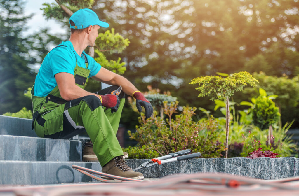 Gardener in green overalls on stone steps, surveying a lush, sunlit garden upgraded by Hook Agency’s hardscaping team and tools.