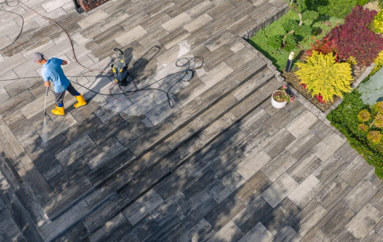 A person in yellow boots power washes a stone patio by vibrant landscaping, clearly demonstrating hardscape cleaning results.