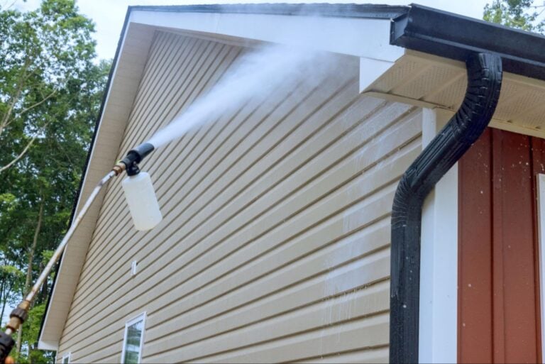 A person power washes beige vinyl siding and white trim, demonstrating industry-approved maintenance for enhanced curb appeal.