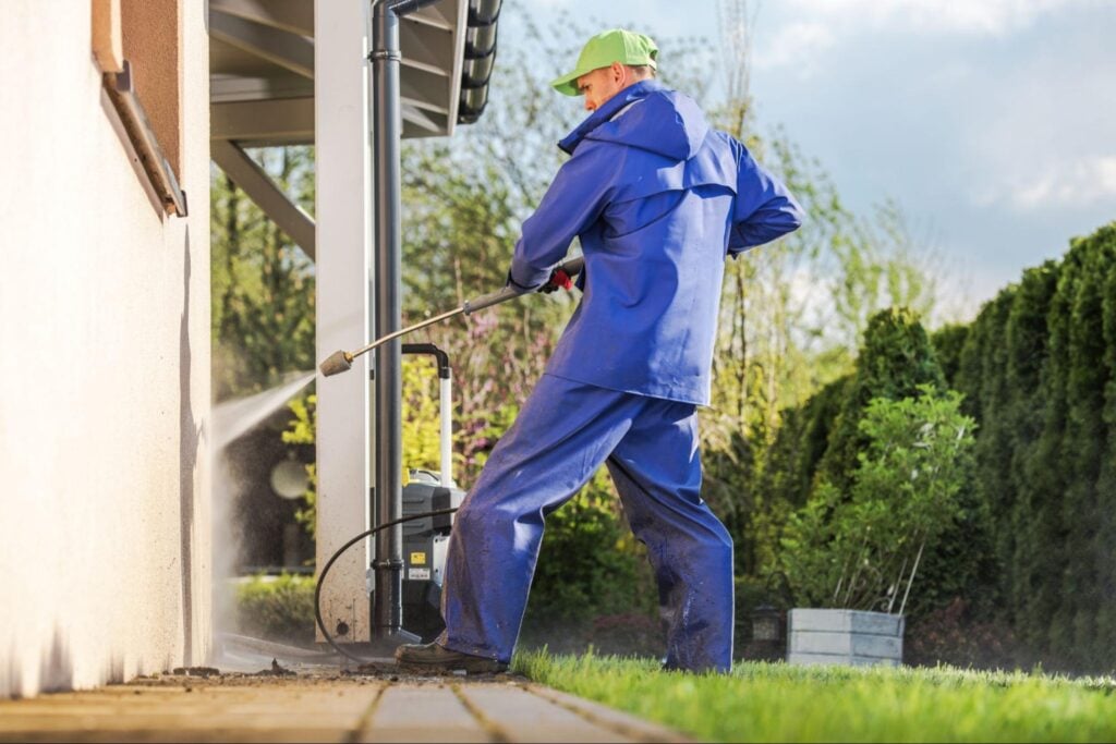 Professional in blue waterproof gear and green cap pressure washes house siding, with foliage behind—ideal for Hook Agency promos.