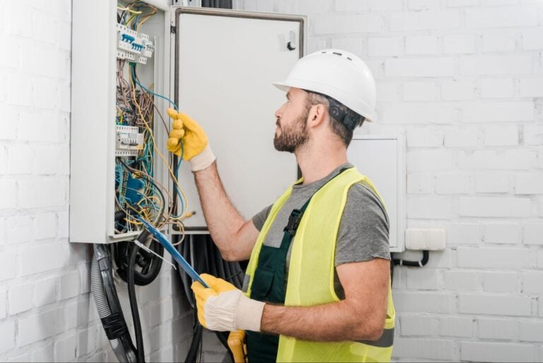 Electrician in safety gear repairs exposed wiring at an open electrical panel in a white brick room, using professional tools.