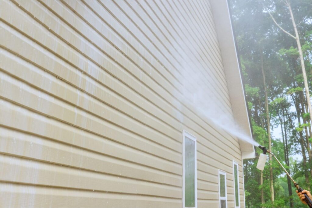 Professional pressure washing removes dirt from beige vinyl siding, demonstrating effective siding cleaning. Trees in background.