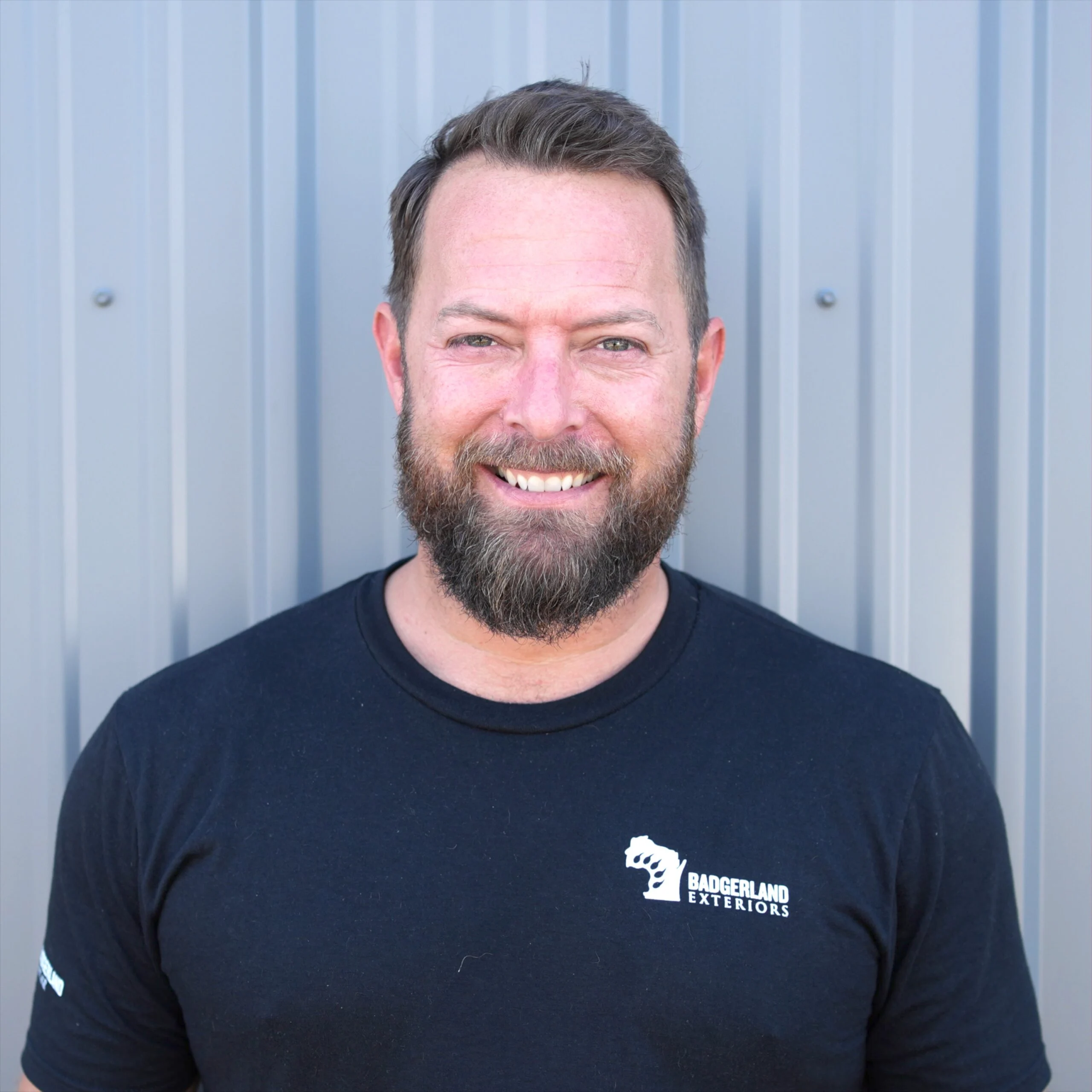 Bearded construction professional in a black "Badgerland Exteriors" T-shirt stands confidently before a corrugated metal wall.