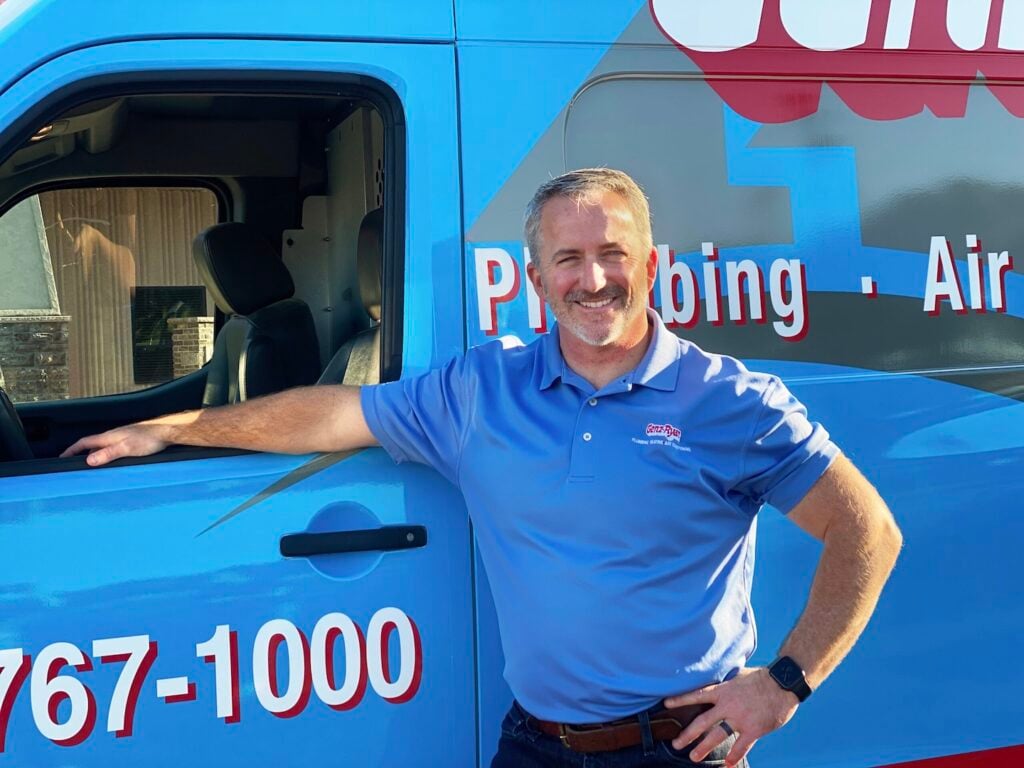 A smiling man in a blue polo stands confidently with one arm on a blue plumbing and air service van featuring red and white branding. The van’s door is open, displaying part of the company phone number.