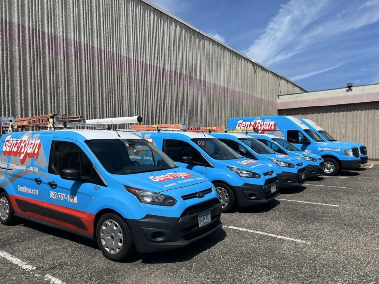 Five professionally branded Genryan service vehicles—blue with red and white logos—are lined up outside a large industrial facility. The fleet includes both vans and trucks equipped with ladders, ready for reliable service.