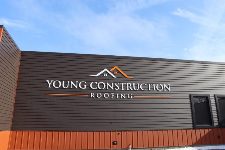 Exterior of a building with dark brown siding and orange trim featuring a prominent "Young Construction Roofing" sign and house roof logo, reflecting trusted construction services—photo for Hook Agency under a blue sky with scattered clouds.
