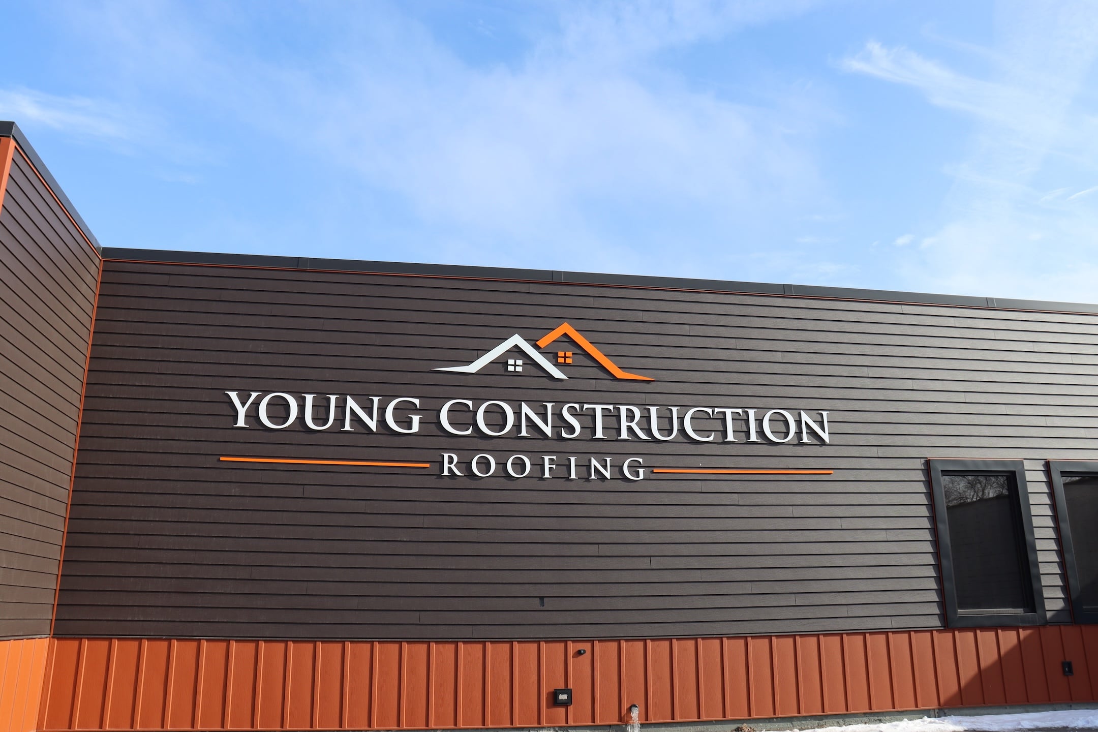 Exterior of a building with dark brown siding and orange trim featuring a prominent "Young Construction Roofing" sign and house roof logo, reflecting trusted construction services—photo for Hook Agency under a blue sky with scattered clouds.