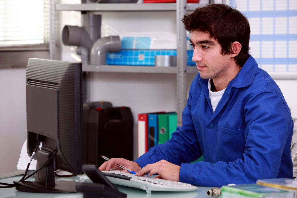 Man in blue lab coat at desk reviews Google Ads data on desktop to reduce cost per lead; shelves and folders in background.