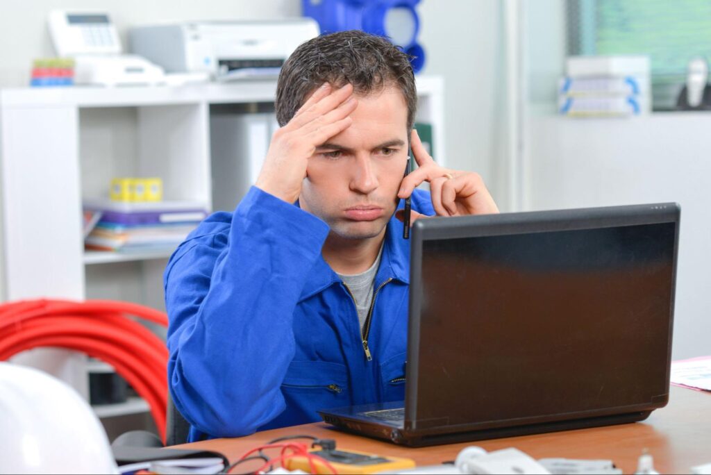 A stressed man in a blue work uniform sits at a desk, on the phone and laptop, worried about home services lead quality.