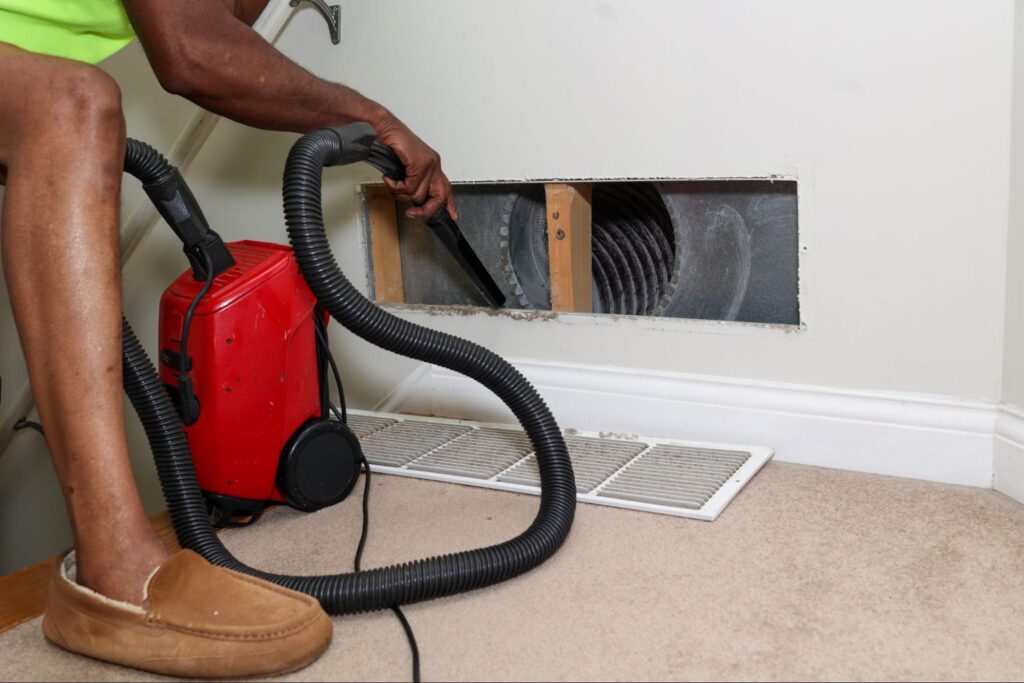 A technician vacuums inside a wall air vent with the cover off—demonstrating a key step for profitable HVAC system maintenance.