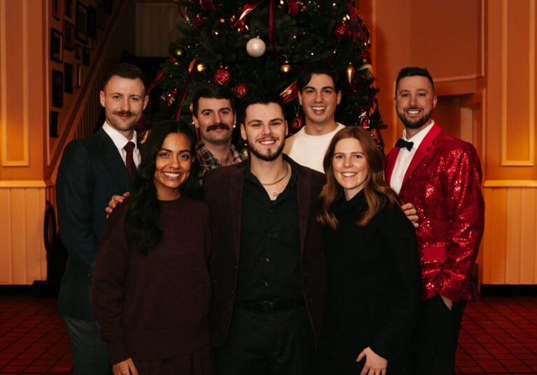 Hook Agency team of seven, formally dressed, smiles by a festive Christmas tree indoors. Four in back row, three in front.