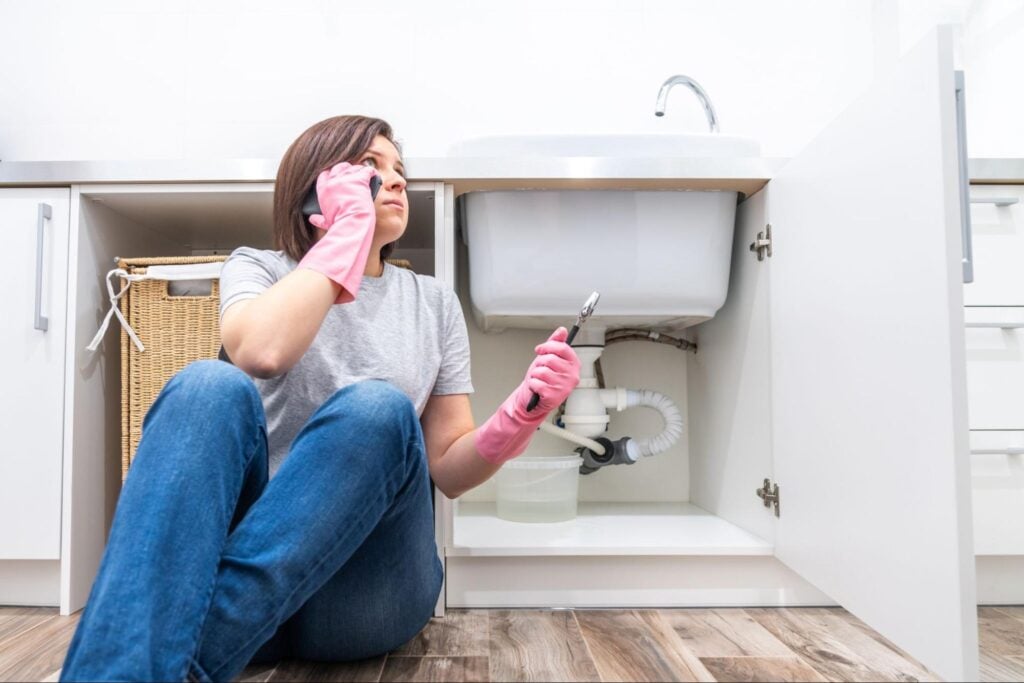 Frustrated woman in pink gloves sits on kitchen floor with wrench and phone, leaking pipe and bucket under sink visible.