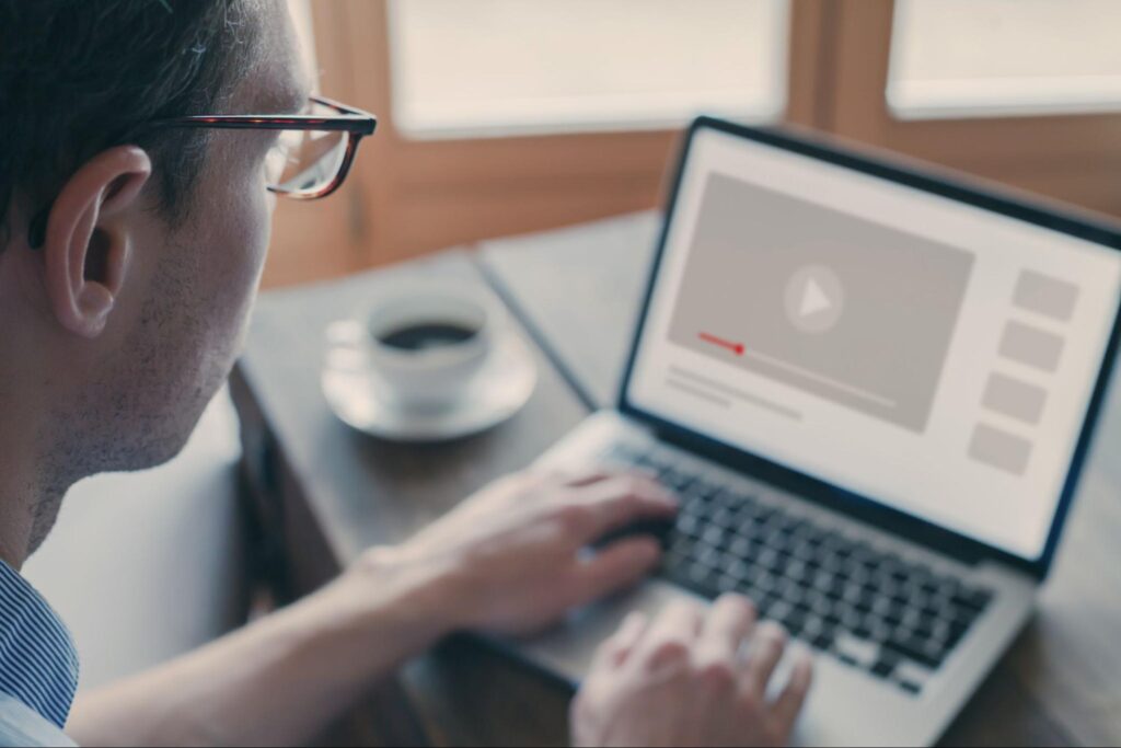 Person with glasses at table, coffee nearby, watches paused Omnipresent Marketing video on laptop; progress bar and thumbnails visible.