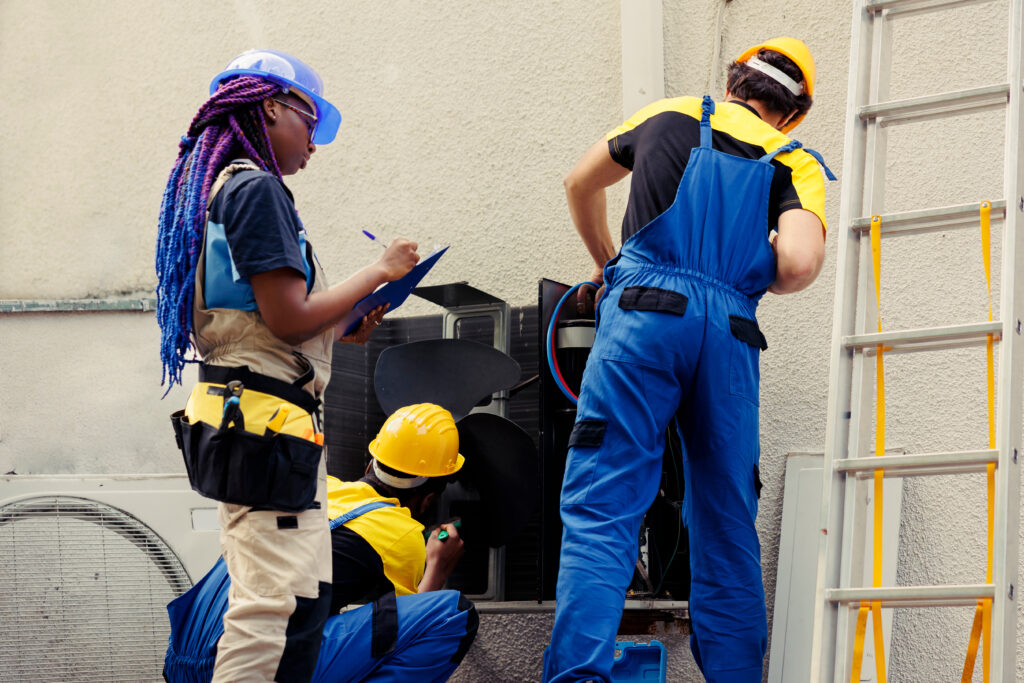 Three HVAC pros in protective gear inspect and service outdoor units. One writes notes; a ladder is positioned nearby for access.