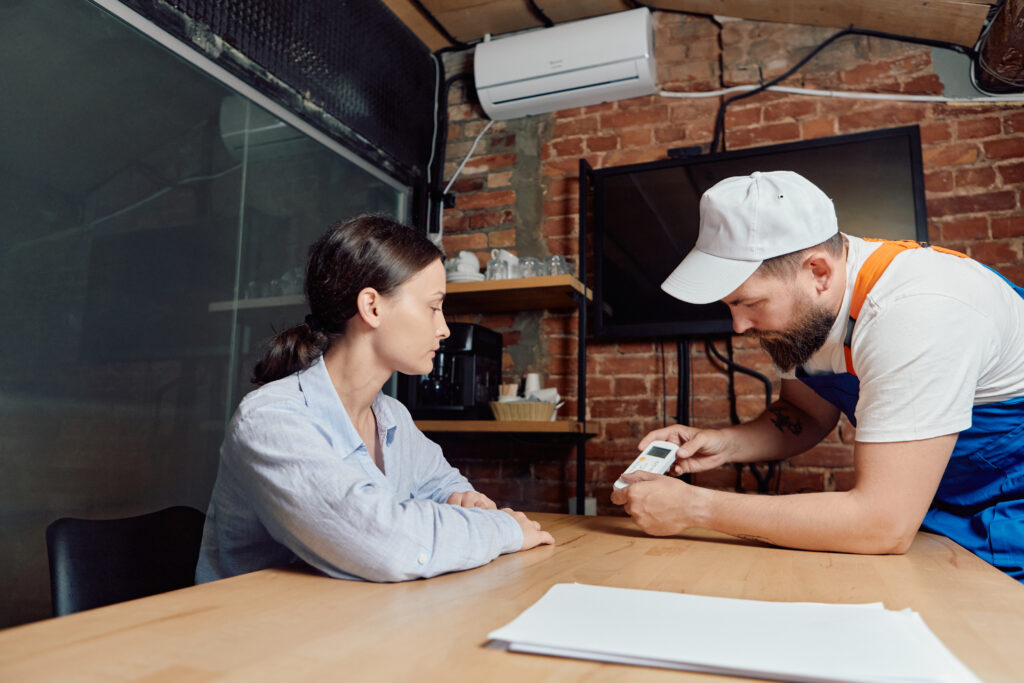 HVAC technician in uniform explains service details on a digital device to a woman at her table, with HVAC unit in background.