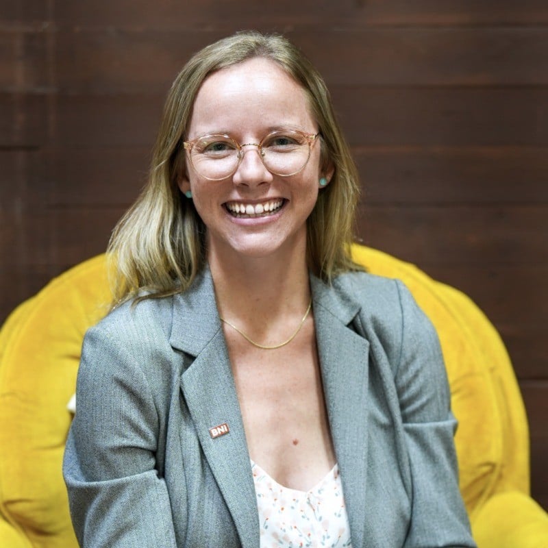 Kristin Sullivan, blonde with glasses, gray blazer, and necklace, smiles on a yellow chair before a wooden wall at Hook Agency.