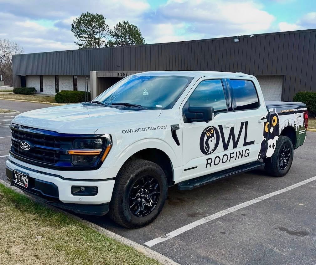 White Ford pickup with black rims, OWL Roofing wrap and owl graphic, website URL on side, parked by a commercial building.