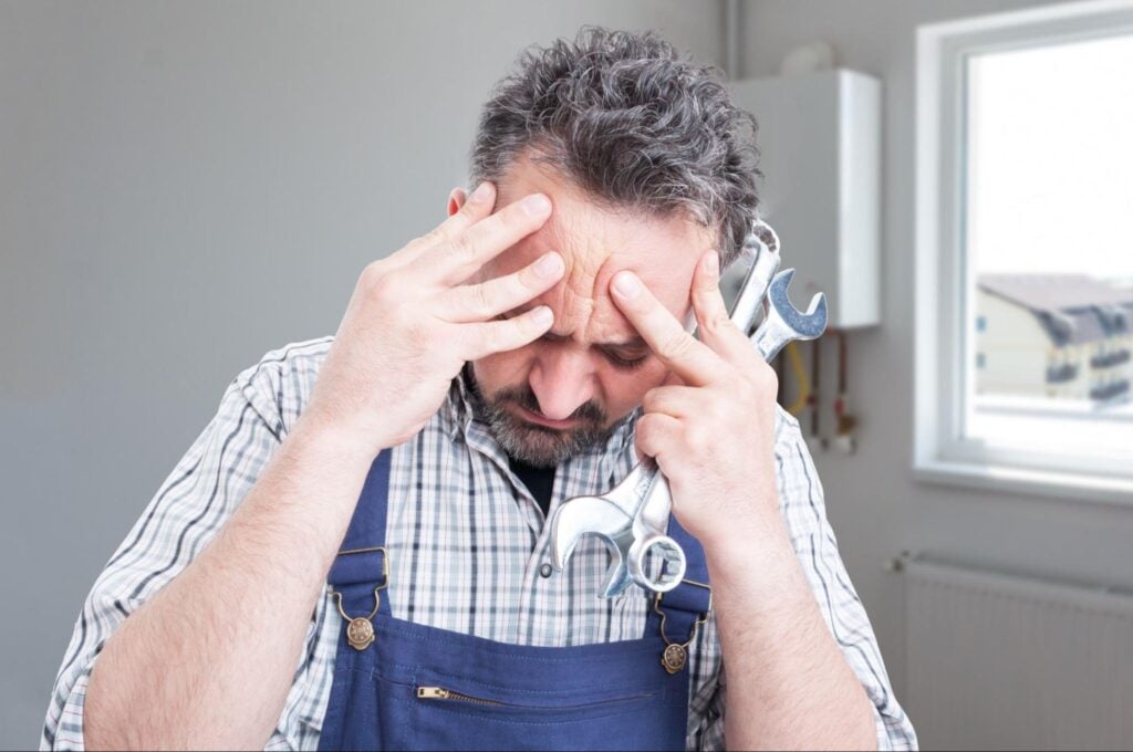 Stressed plumber in overalls with wrenches, pressing fingers to forehead indoors—overwhelmed by sales challenges.