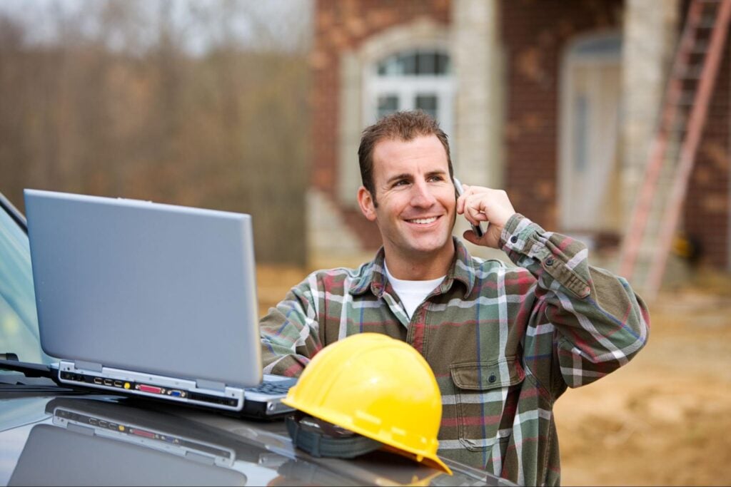 Contractor in plaid shirt discusses roofing AI on phone at job site, with laptop and hard hat on vehicle; building in background.