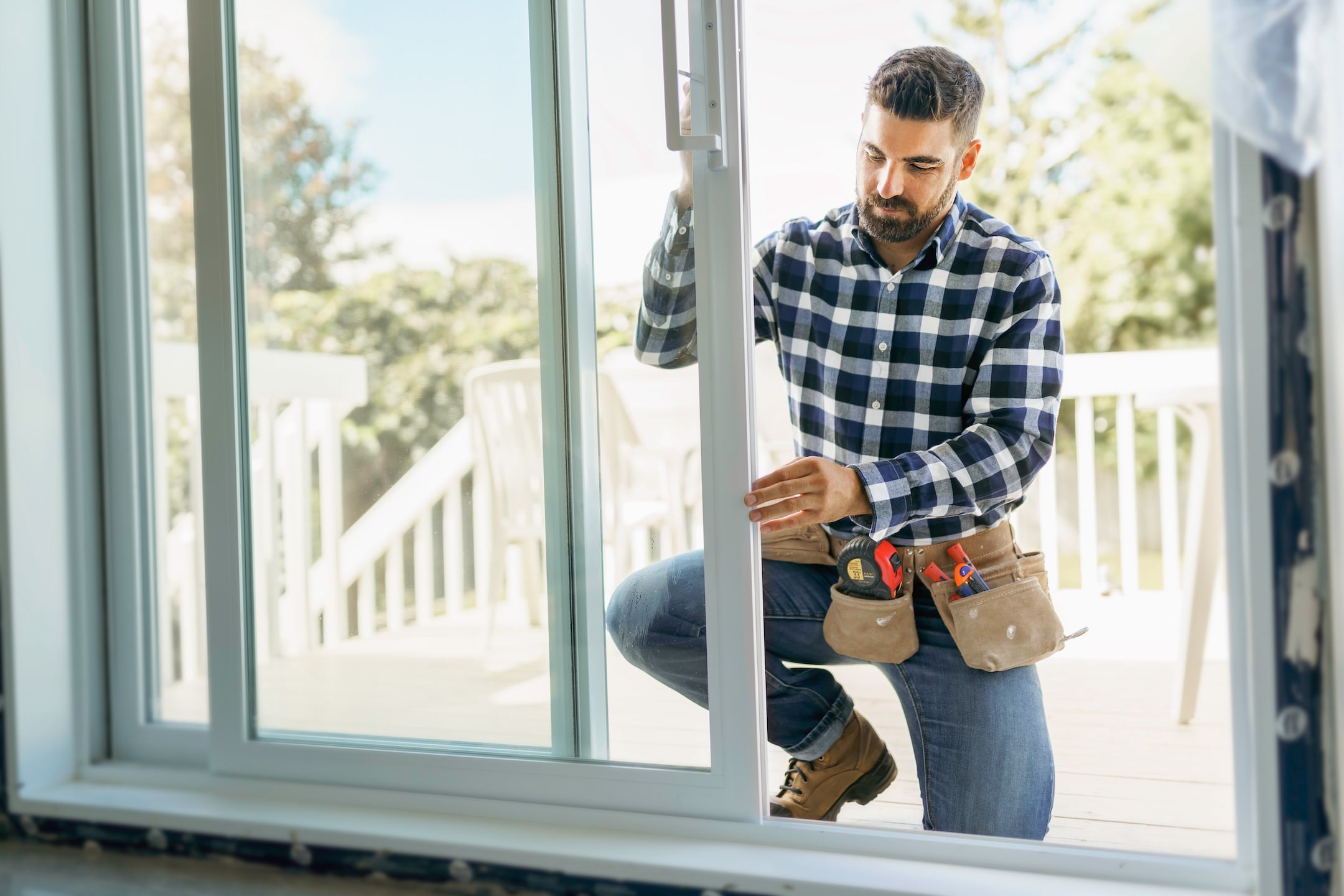 Professional in plaid shirt and tool belt kneels to adjust sliding glass door, with outdoor deck and trees in the background.
